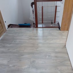 Sucha zabudowa , szpachlowanie , malowanie , podłogi, laminat, winyl - Jasnoszara podłoga panelowa ułożona w korytarzu, widoczne drzwi z naturalnego drewna i fragment schodów z drewnianą balustradą ze stalowymi prętami.