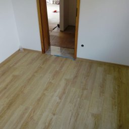 Sucha zabudowa , szpachlowanie , malowanie , podłogi, laminat, winyl - Pokój po remoncie z nową podłogą imitującą drewno i świeżo pomalowanymi białymi ścianami, widoczne zabezpieczenie folią na progu w przejściu do kolejnego pomieszczenia.