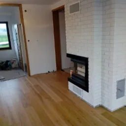 Wnętrze w trakcie wykańczania: kominek obłożony białą cegłą, drewniana podłoga, widoczne niedokończone elementy i okablowanie.