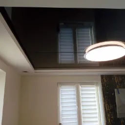 Czarne, lustrzane wykończenie sufitu z widocznym odbiciem okna i lampy, w tle ściana z dekoracyjną tapetą.