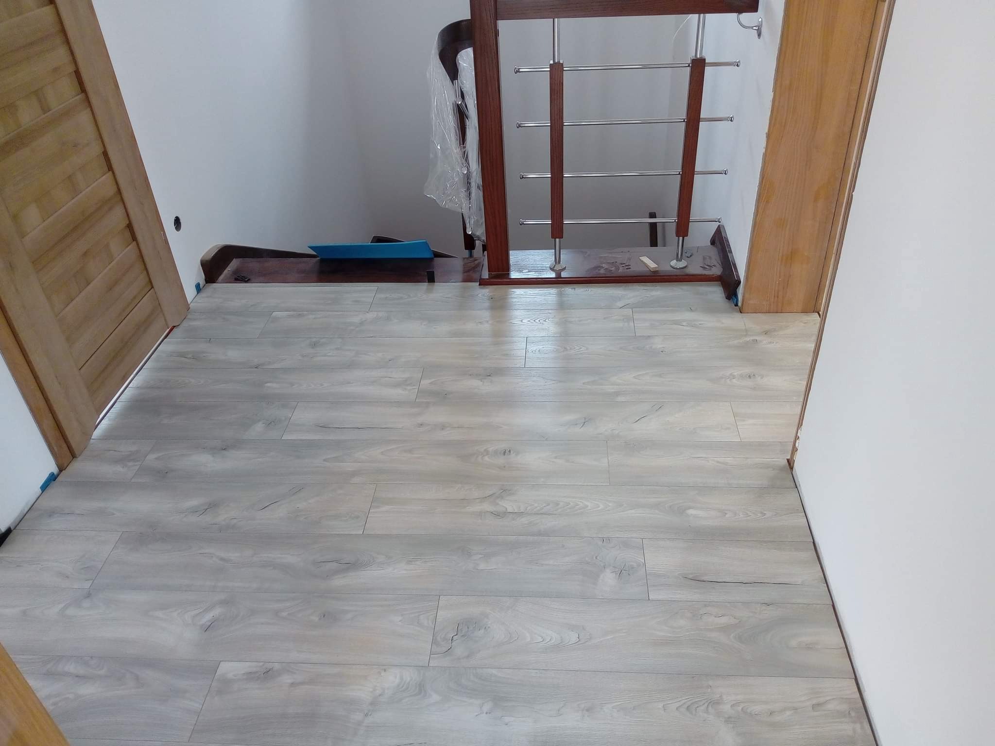 Jasnoszara podłoga panelowa ułożona w korytarzu, widoczne drzwi z naturalnego drewna i fragment schodów z drewnianą balustradą ze stalowymi prętami.