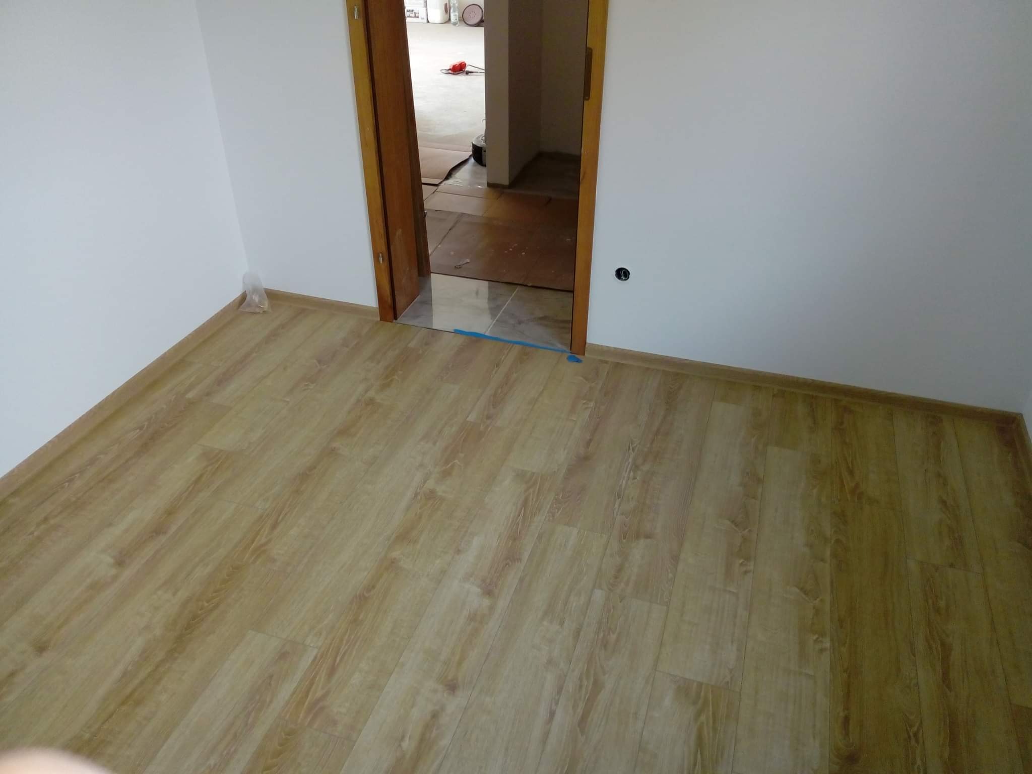 Pokój po remoncie z nową podłogą imitującą drewno i świeżo pomalowanymi białymi ścianami, widoczne zabezpieczenie folią na progu w przejściu do kolejnego pomieszczenia.