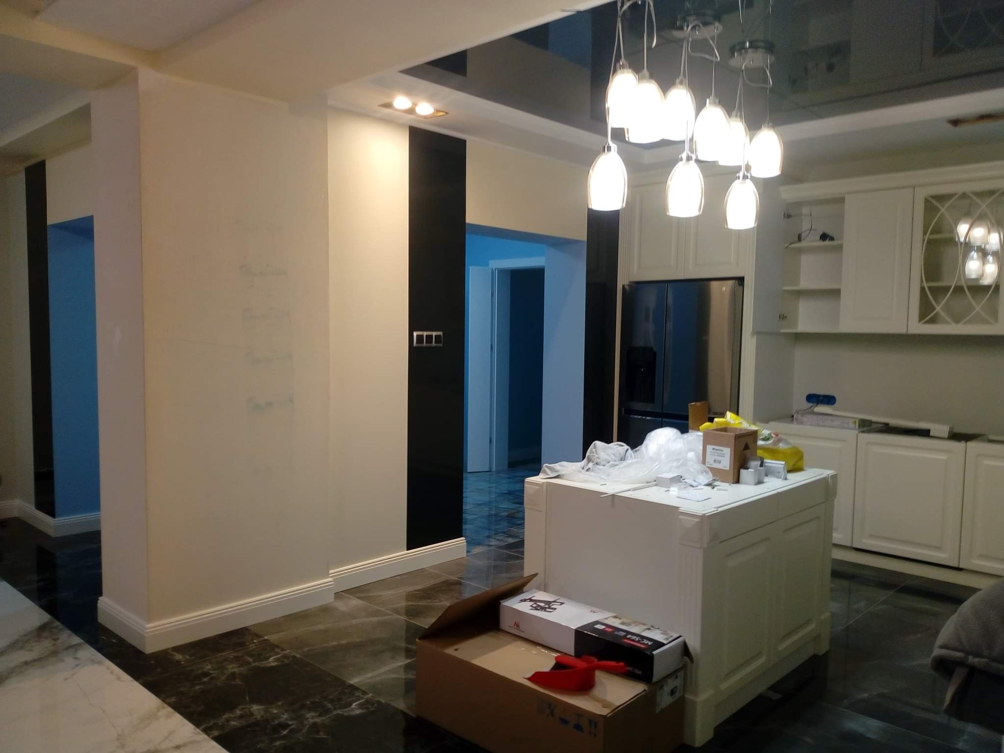 Wnętrze w trakcie remontu: białe ściany z czarnymi akcentami, błyszczący czarny sufit z nowoczesnym oświetleniem, marmurowa podłoga, kartony na wyspie kuchennej, widoczne przygotowania do prac...