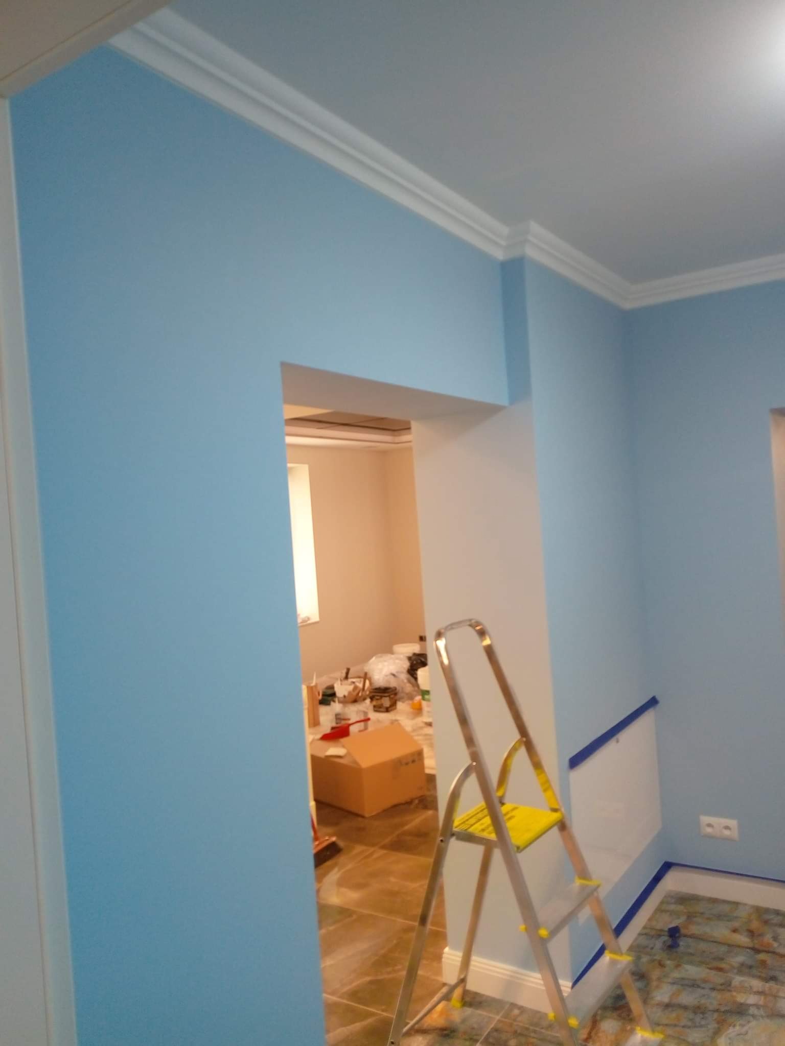 Świeżo pomalowane na błękitno ściany z białym sztukateryjnym wykończeniem pod sufitem, widoczna drabina malarska i otwarte przejście do pomieszczenia z materiałami budowlanymi.