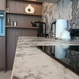 BPM Bujanowski Pracownia Mebli - Fragment nowoczesnej kuchni z blatem imitującym marmur, czarną armaturą i płytą indukcyjną, na półkach dekoracje, w tle ściana z wzorem marmuru.