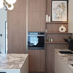 BPM Bujanowski Pracownia Mebli - Nowoczesna kuchnia z drewnianymi frontami szafek, marmurowym blatem wyspy i białymi szafkami dolnymi, uzupełniona o wiszące półki z dekoracjami, oświetlona lampą w stylu art deco.