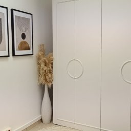 BPM Bujanowski Pracownia Mebli - Biała szafa na wymiar z okrągłymi uchwytami, minimalistyczne obrazy w czarnych ramach i dekoracyjna trawa pampasowa w białym wazonie.
