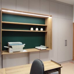 BPM Bujanowski Pracownia Mebli - Biuro w Olsztynie z zabudową meblową: szafy w kolorze szarym, drewniane półki z oświetleniem LED i zielonym tłem, biurko z dokumentami, drukarka, otwarte drzwi.