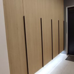 BPM Bujanowski Pracownia Mebli - Długa szafa wnękowa z jasnego drewna z pionowymi czarnymi uchwytami, podświetlona od dołu, na tle białych płytek podłogowych i czarnych drzwi.
