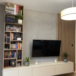 BPM Bujanowski Pracownia Mebli - Nowoczesny salon z telewizorem, białą szafką RTV, regałem z książkami i grami, oraz dekoracyjną ścianką z drewnianych listew.