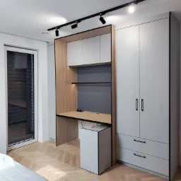 Nowoczesny pokój z zabudową meblową: szafa, biurko z półkami i szafka pomocnicza w odcieniach szarości i drewna, oświetlenie punktowe na suficie, parkiet w jodełkę.