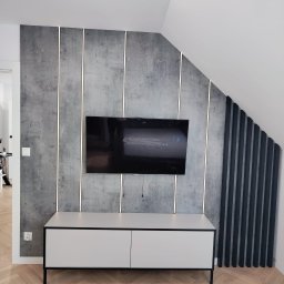 BPM Bujanowski Pracownia Mebli - Telewizor zawieszony na ścianie z betonową fakturą, podświetlonej pionowymi paskami LED, z białą szafką RTV na czarnych nóżkach i drewnianą podłogą ułożoną w jodełkę.