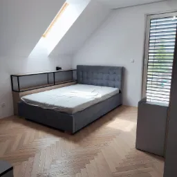 Sypialnia na poddaszu z szarym łóżkiem z zagłówkiem, drewnianą zabudową z półkami nad łóżkiem i parkietem w jodełkę.