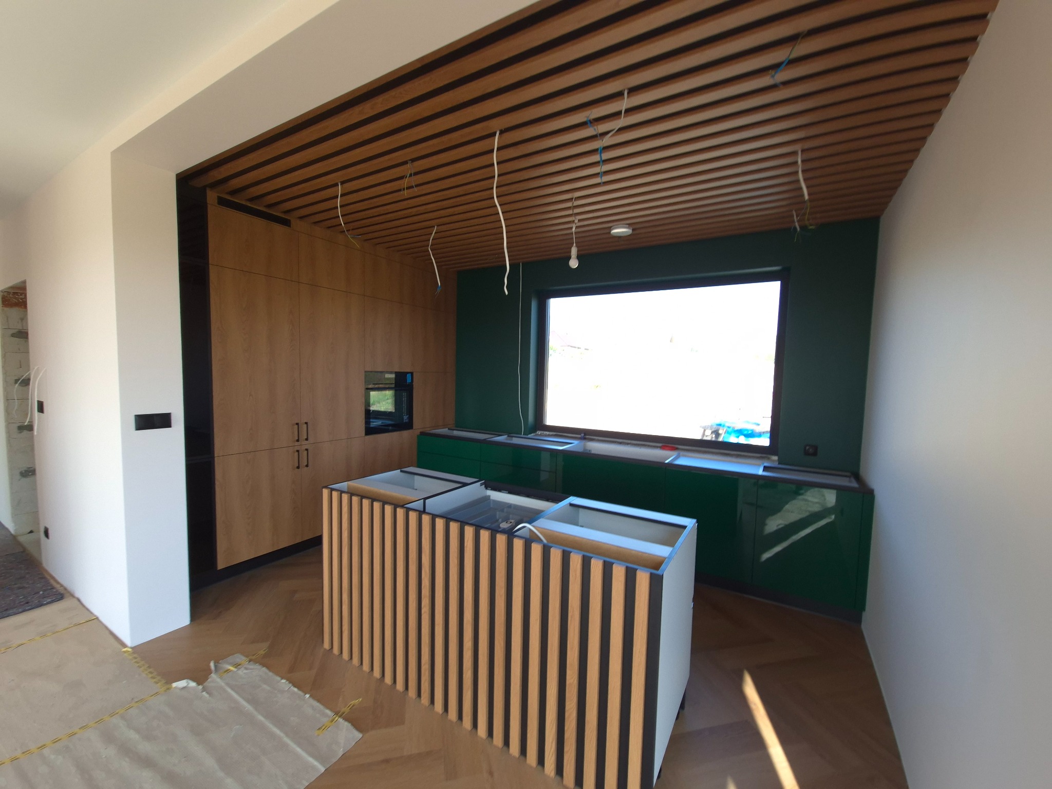 Wnętrze kuchni w trakcie montażu: drewniane szafki, zielone ściany, wyspa z drewnianymi lamelami, widoczne przewody elektryczne zwisające z sufitu z drewnianą konstrukcją.
