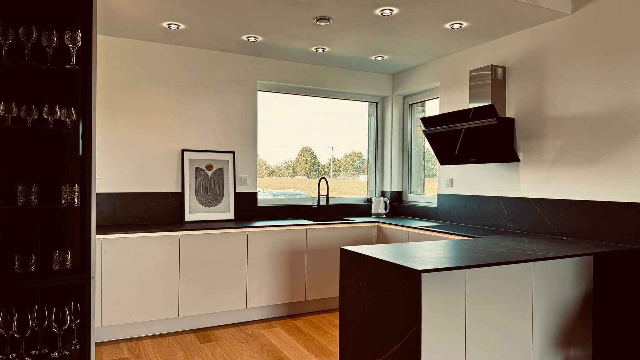 Nowoczesna kuchnia z jasnymi frontami szafek, czarnym blatem z marmurowym wzorem, drewnianą podłogą i oknem z widokiem na zieleń, oświetlona punktowymi lampami sufitowymi.