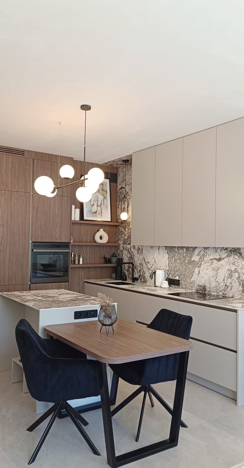 Nowoczesna kuchnia z wyspą i stołem jadalnym, drewniane fronty szafek połączone z beżowymi, marmurowy backsplash, oświetlenie punktowe i wiszące lampy kuliste.