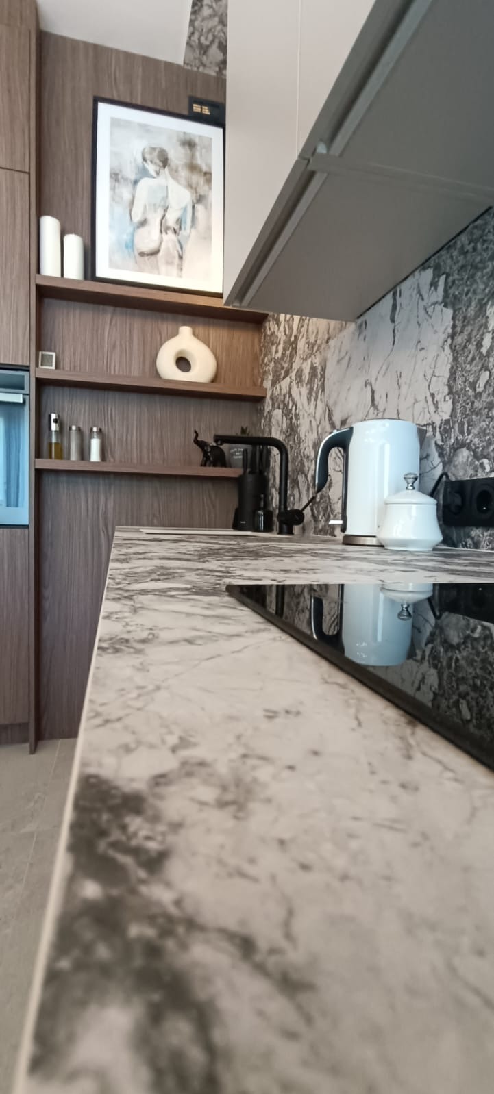 Fragment nowoczesnej kuchni z blatem imitującym marmur, czarną armaturą i płytą indukcyjną, na półkach dekoracje, w tle ściana z wzorem marmuru.