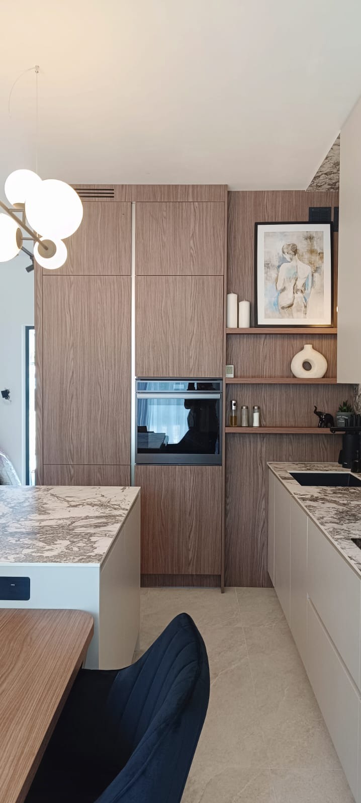 Nowoczesna kuchnia z drewnianymi frontami szafek, marmurowym blatem wyspy i białymi szafkami dolnymi, uzupełniona o wiszące półki z dekoracjami, oświetlona lampą w stylu art deco.