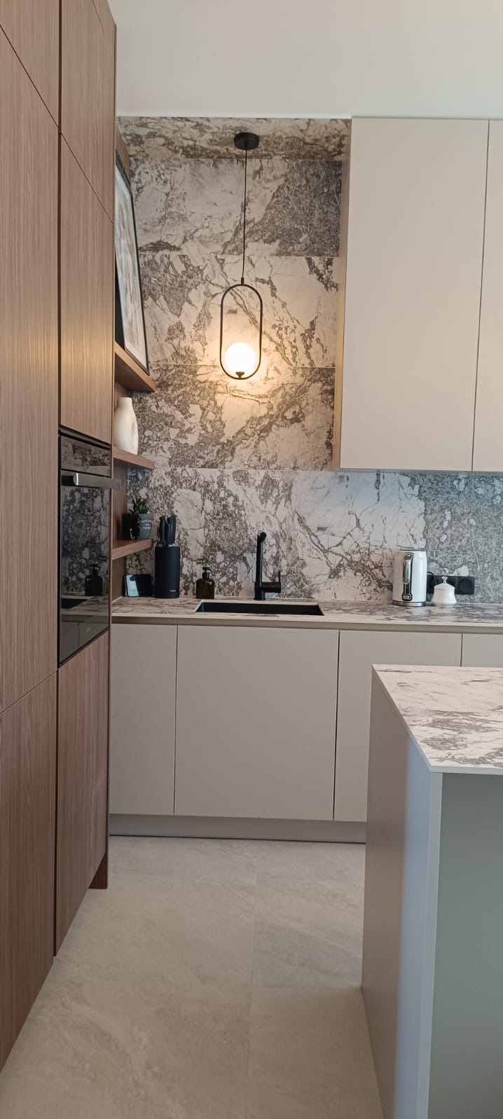 Nowoczesna kuchnia z drewnianymi szafkami, blatem imitującym marmur, czarną armaturą i wiszącą lampą o owalnym kształcie, oświetlającą przestrzeń roboczą