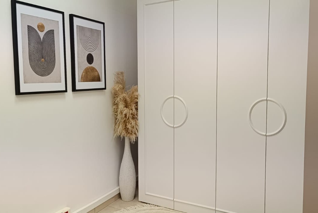 Biała szafa na wymiar z okrągłymi uchwytami, minimalistyczne obrazy w czarnych ramach i dekoracyjna trawa pampasowa w białym wazonie.