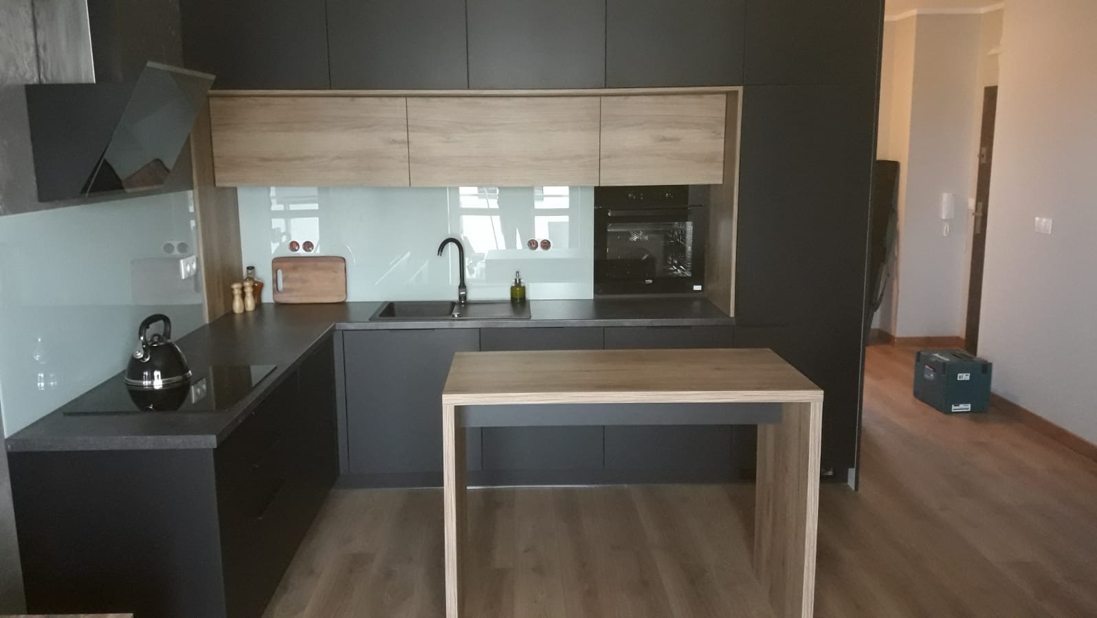 Nowoczesna kuchnia z ciemnymi szafkami i drewnianymi akcentami, wyspa kuchenna z drewnianym blatem, widoczna skrzynka narzędziowa w tle.