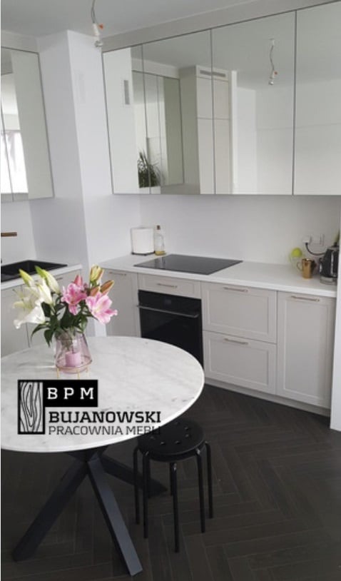 Jasna kuchnia z białymi szafkami wiszącymi z lustrzanymi frontami, okrągły marmurowy stół z bukietem lilii w wazonie, czarny stołek i parkiet ułożony w jodełkę.