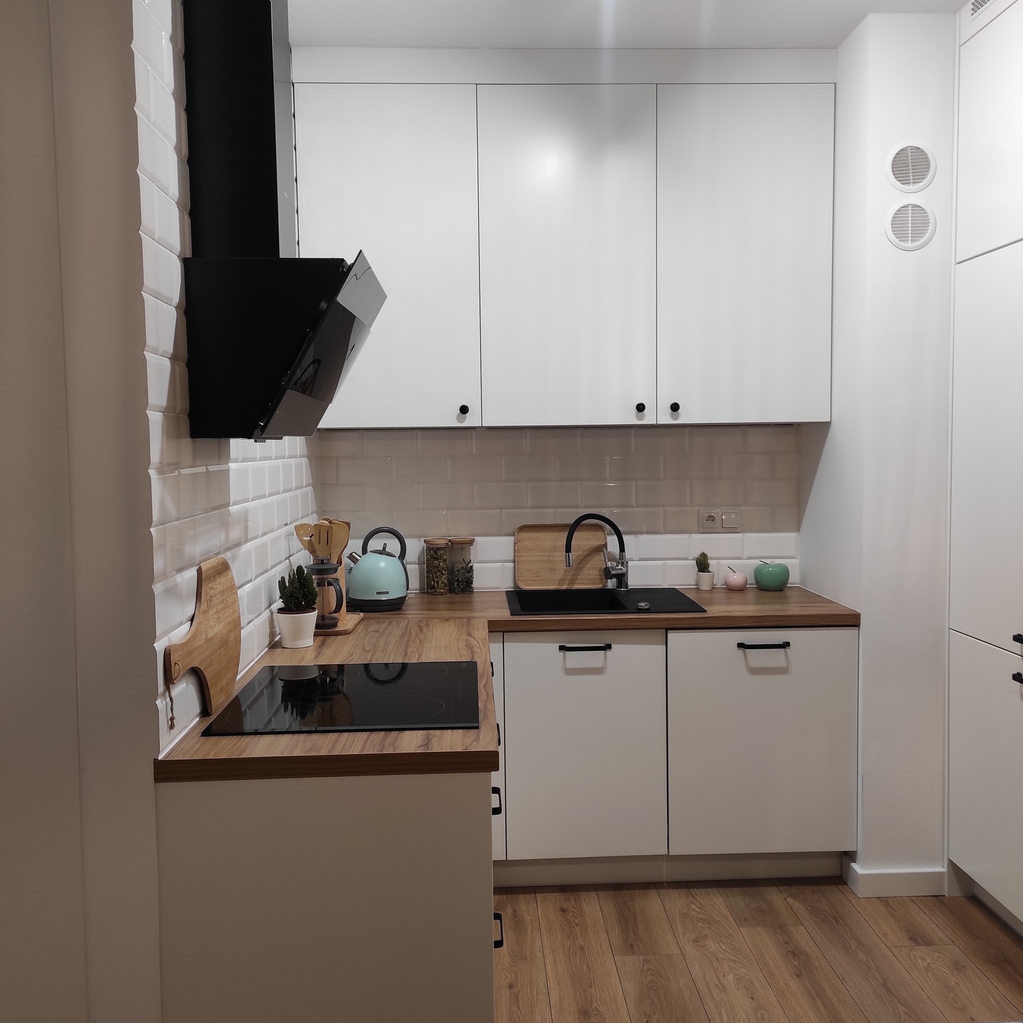 Nowoczesna, biała kuchnia z drewnianym blatem, czarną płytą indukcyjną i okapem. Na blacie czajnik w kolorze pastelowej zieleni, akcesoria kuchenne i małe rośliny w doniczkach.