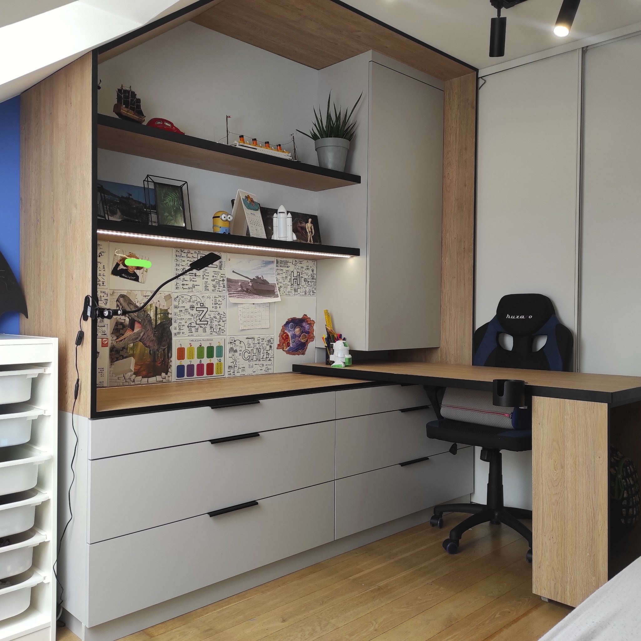 Pokój młodzieżowy z biurkiem w zabudowie, szare fronty szafek z czarnymi uchwytami, drewniane elementy wykończeniowe, oświetlenie LED pod półkami, na ścianie przy biurku rysunki i zdjęcia.