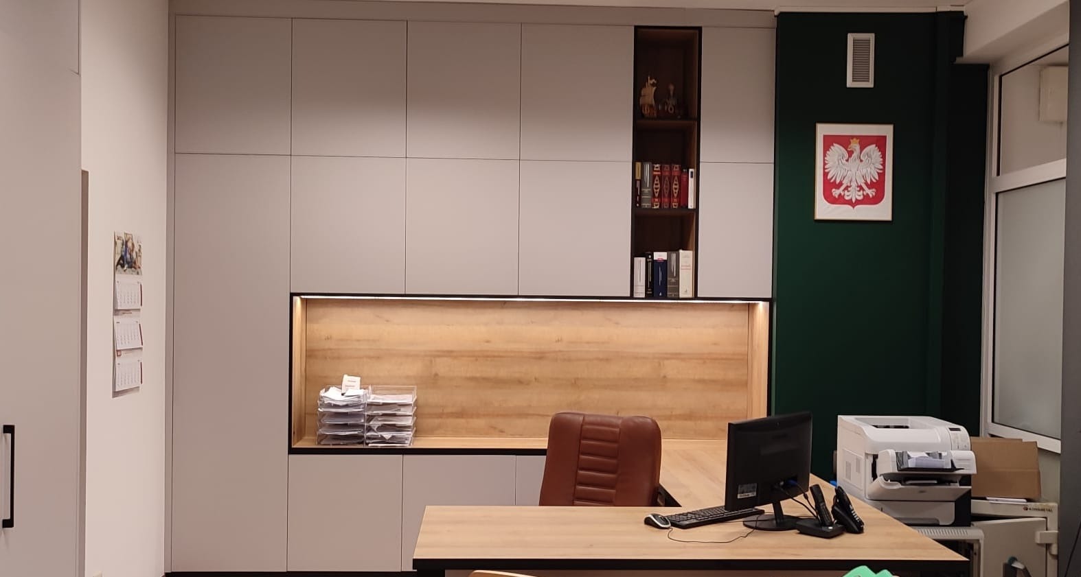 Biuro z szarymi szafami na wymiar z drewnianymi elementami, biurkiem z komputerem i fotelem, na ścianie godło Polski.