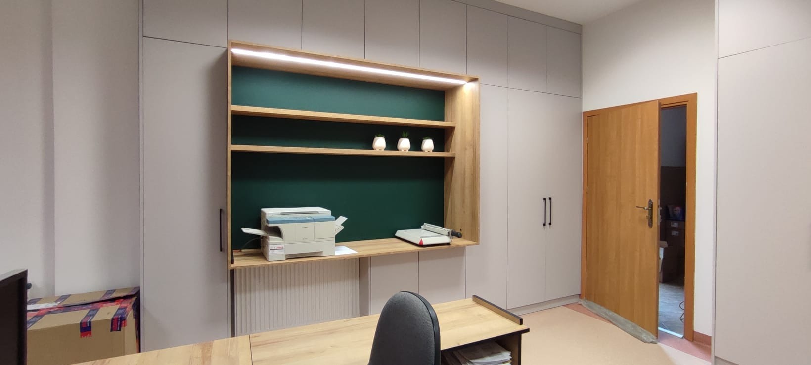 Biuro w Olsztynie z zabudową meblową: szafy w kolorze szarym, drewniane półki z oświetleniem LED i zielonym tłem, biurko z dokumentami, drukarka, otwarte drzwi.