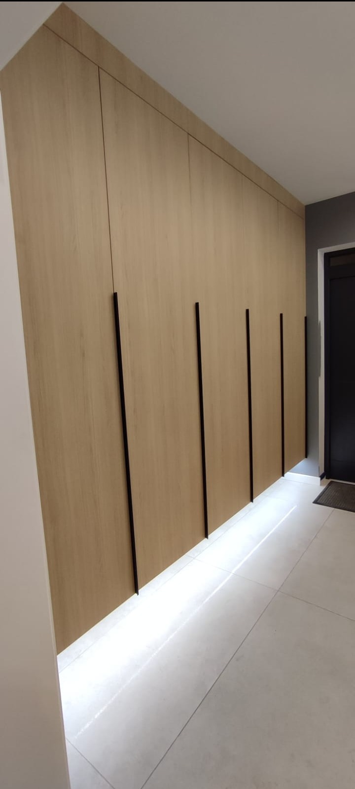 Długa szafa wnękowa z jasnego drewna z pionowymi czarnymi uchwytami, podświetlona od dołu, na tle białych płytek podłogowych i czarnych drzwi.