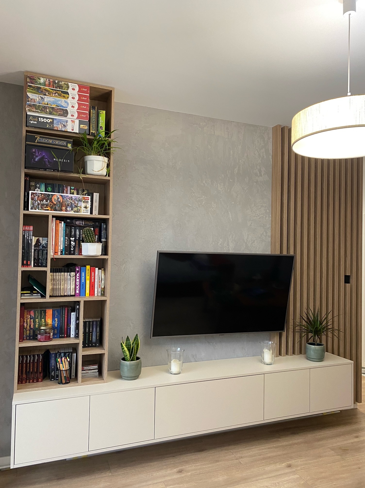 Nowoczesny salon z telewizorem, białą szafką RTV, regałem z książkami i grami, oraz dekoracyjną ścianką z drewnianych listew.