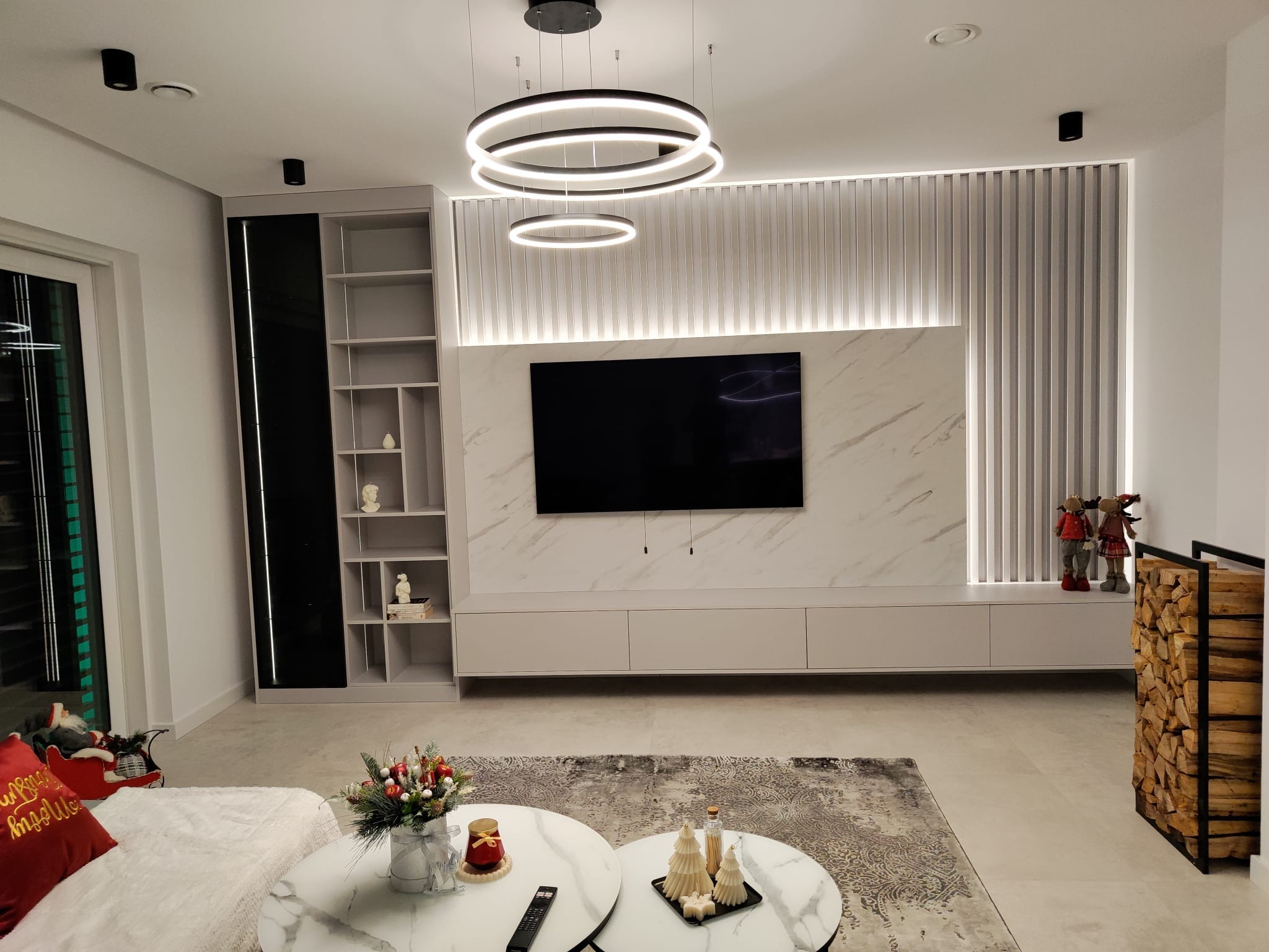 Nowoczesny salon z marmurową ścianą TV, podświetlanymi lamelami, okrągłymi lampami i dekoracjami świątecznymi na stolikach kawowych.