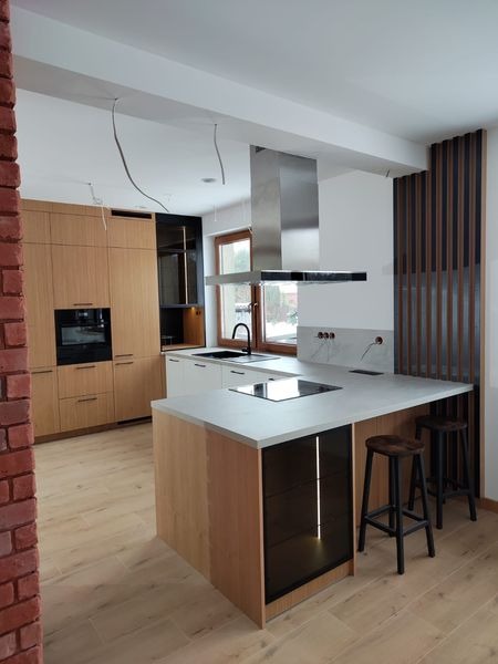 Jasna kuchnia z drewnianymi frontami, wyspą z szarym blatem i czarnymi akcentami, widoczne niedokończone podłączenia elektryczne.