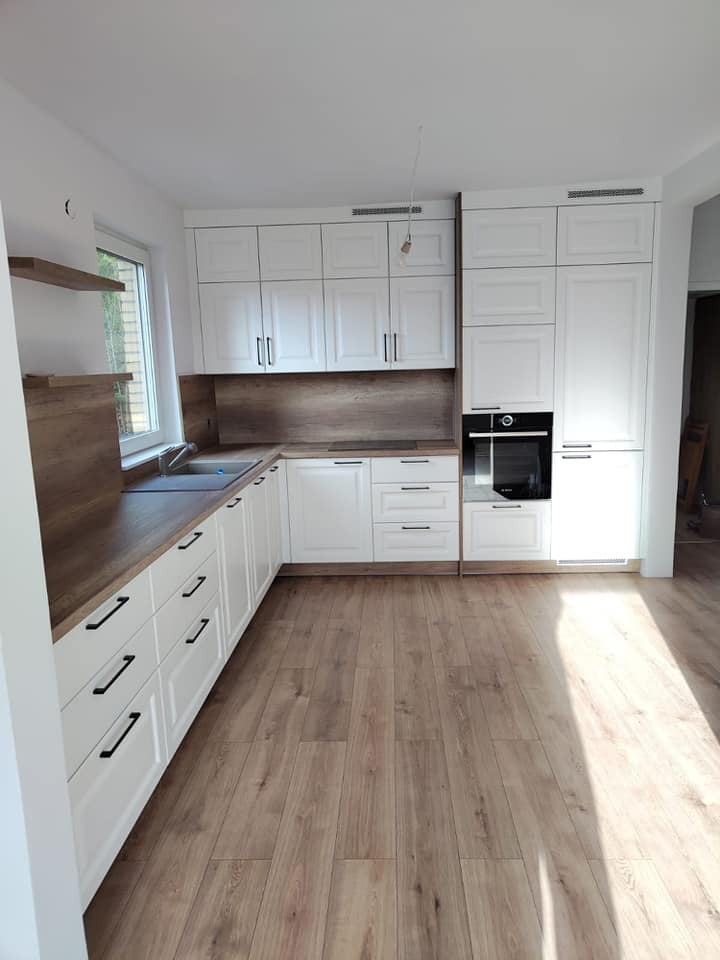 Nowoczesna kuchnia z białymi frontami szafek i drewnianymi blatami, czarnymi uchwytami, drewniana podłoga, piekarnik w zabudowie, widok z perspektywy podłogi.