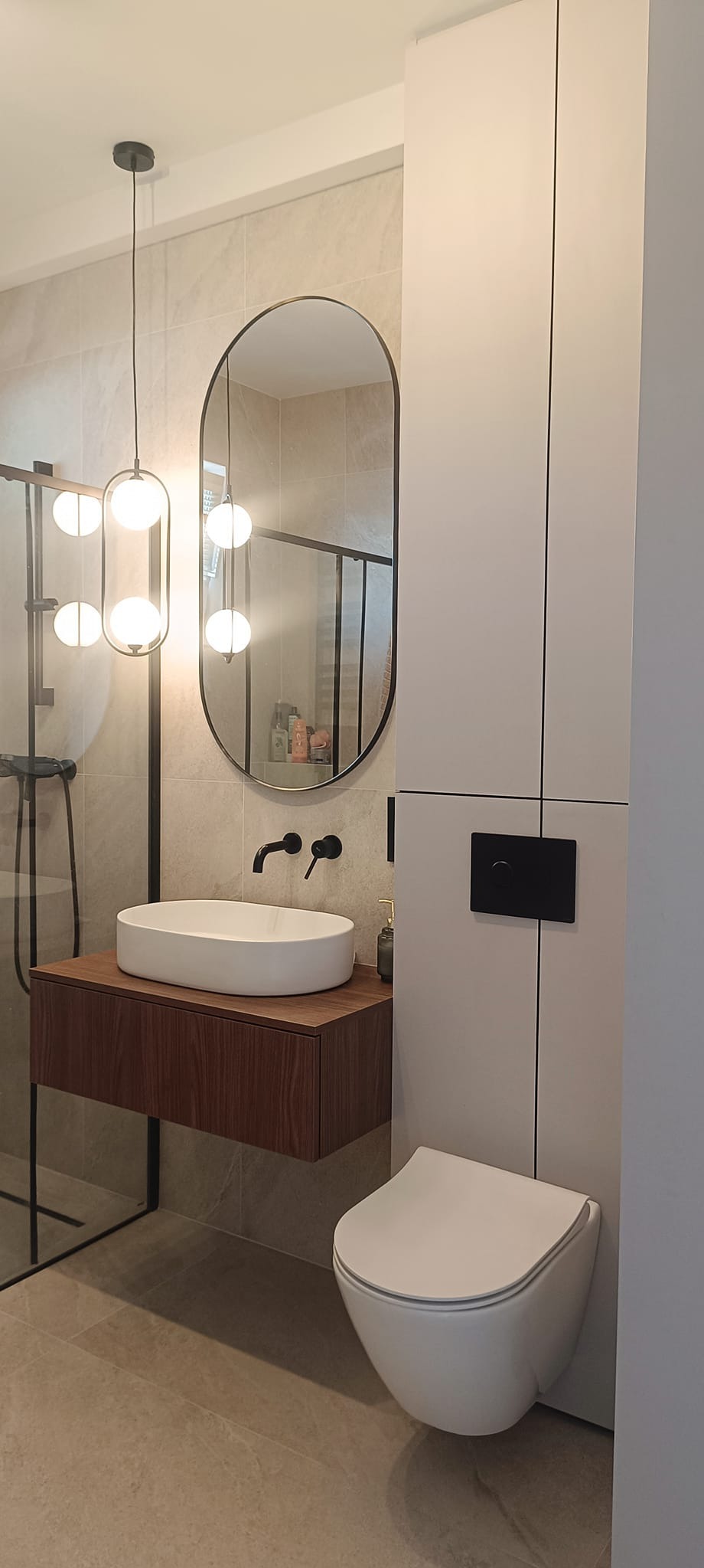 Nowoczesna łazienka z owalną umywalką nablatową na drewnianej szafce, wisząca toaleta i podświetlane lustro owalne, oświetlenie kuliste, armatura w kolorze czarnym, beżowe płytki.