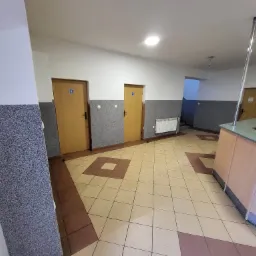 Wnętrze korytarza z dwoma drzwiami oznaczonymi ikoną toalety, grzejnikiem, wykończeniem ścian w szarym melanżu i płytkami podłogowymi w odcieniach beżu i brązu.