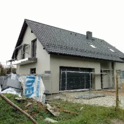 Nowoczesny dom jednorodzinny w trakcie ocieplania ścian zewnętrznych, widoczne rusztowania, materiały izolacyjne i świeżo otynkowana elewacja w kolorze szarym.