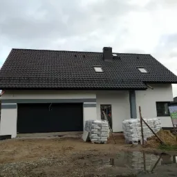 Nowo wybudowany dom jednorodzinny z ciemną dachówką i szarą stolarką, widoczne worki z materiałami budowlanymi na posesji, po deszczu.