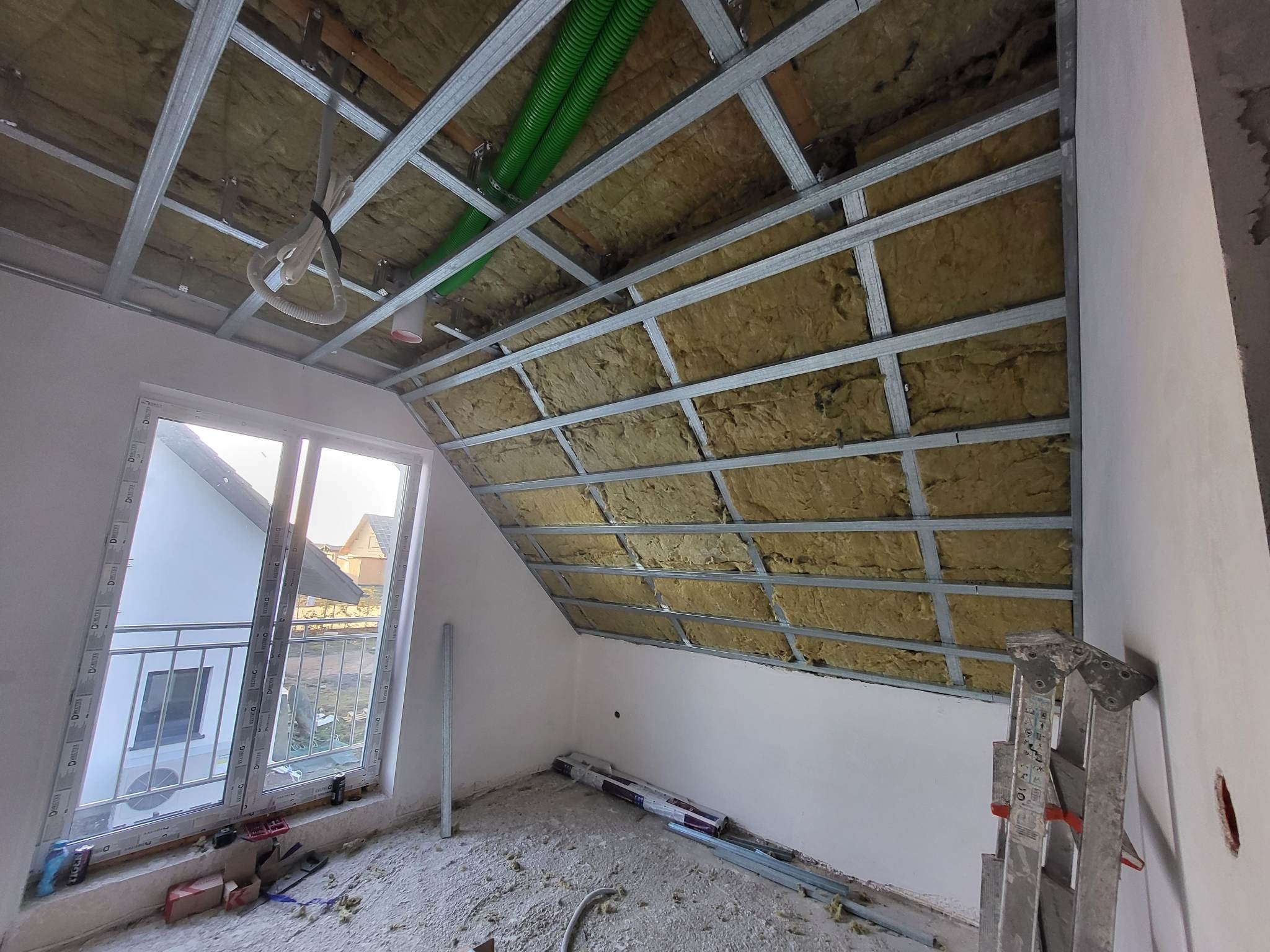 Poddasze w trakcie prac wykończeniowych: widoczna izolacja termiczna między metalowymi profilami sufitowymi, zielone rury wentylacyjne, okno dachowe z widokiem na dom, drabina oparta o ścianę...