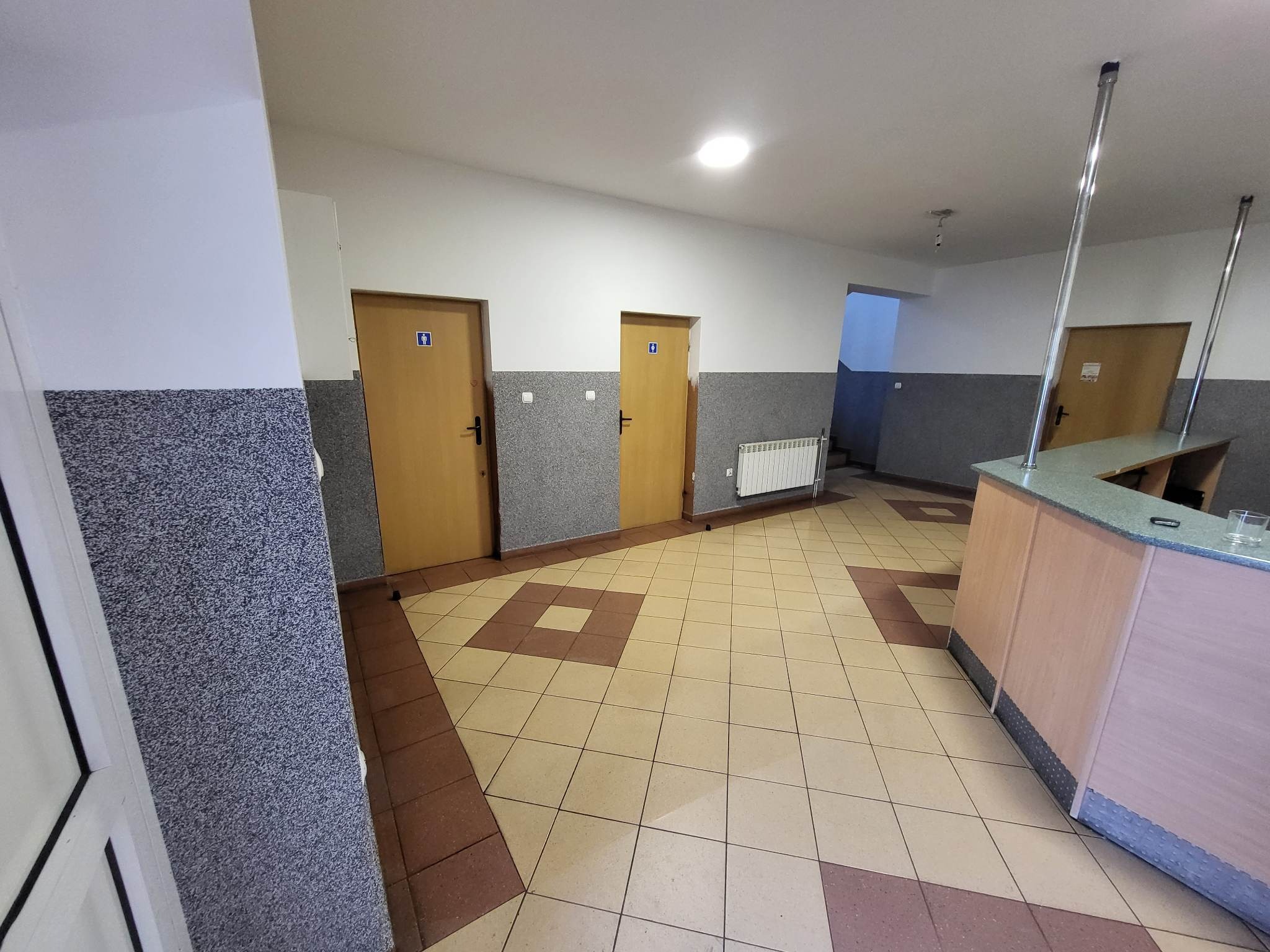 Wnętrze korytarza z dwoma drzwiami oznaczonymi ikoną toalety, grzejnikiem, wykończeniem ścian w szarym melanżu i płytkami podłogowymi w odcieniach beżu i brązu.