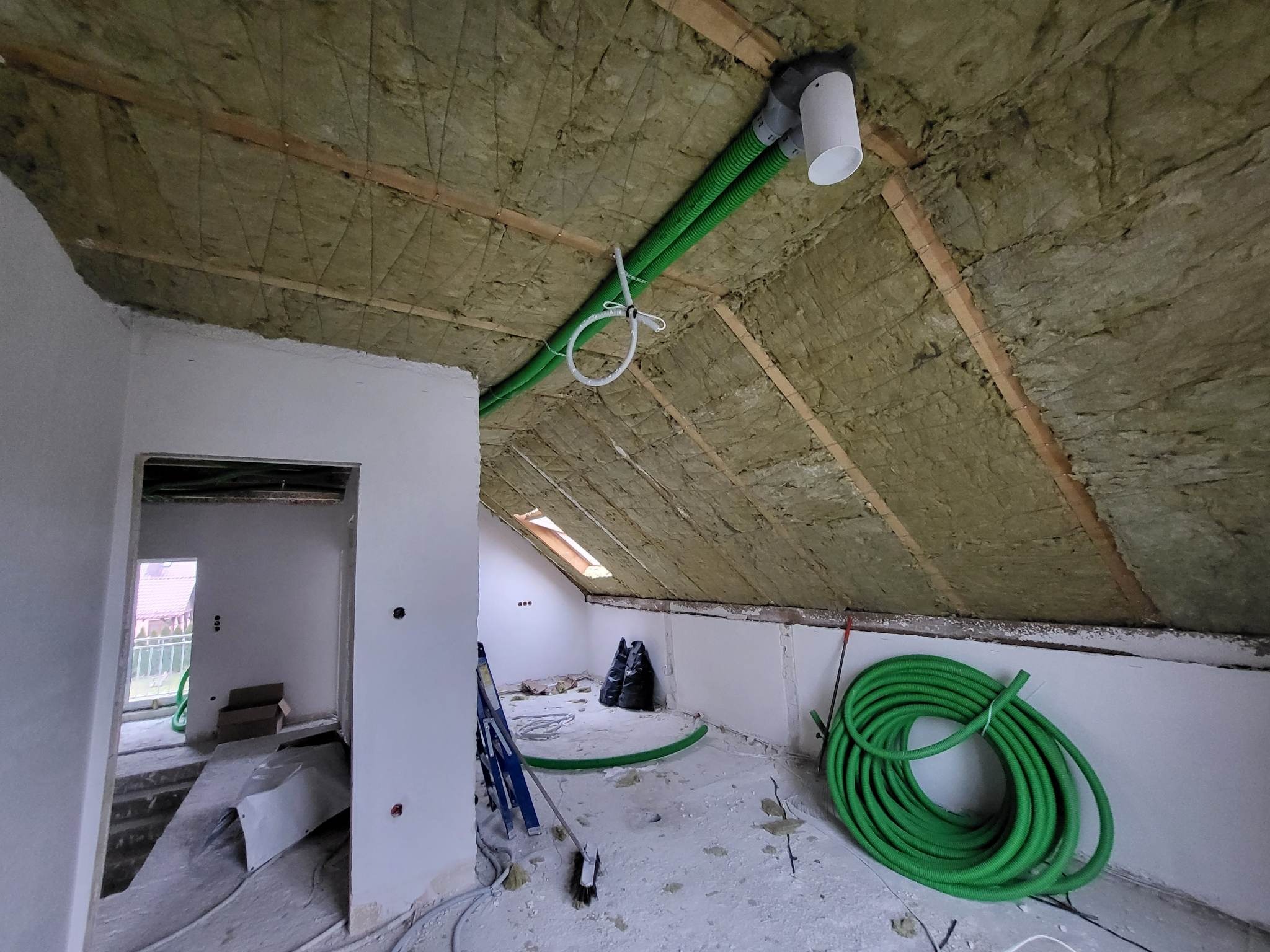 Poddasze w trakcie prac, widoczna izolacja z wełny mineralnej na skośnym dachu, zielone rury wentylacyjne, drabina oparta o ścianę i okno dachowe.
