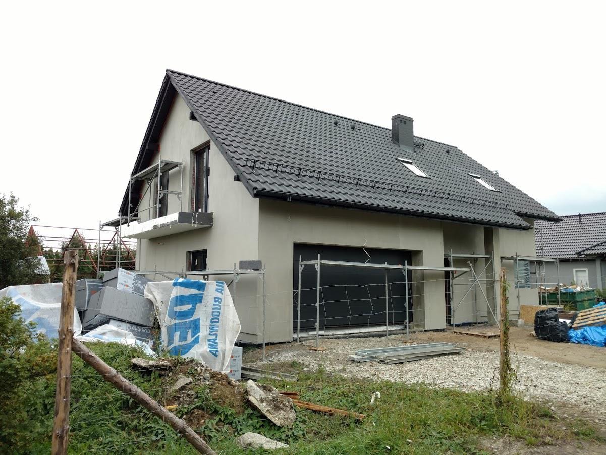 Nowoczesny dom jednorodzinny w trakcie ocieplania ścian zewnętrznych, widoczne rusztowania, materiały izolacyjne i świeżo otynkowana elewacja w kolorze szarym.