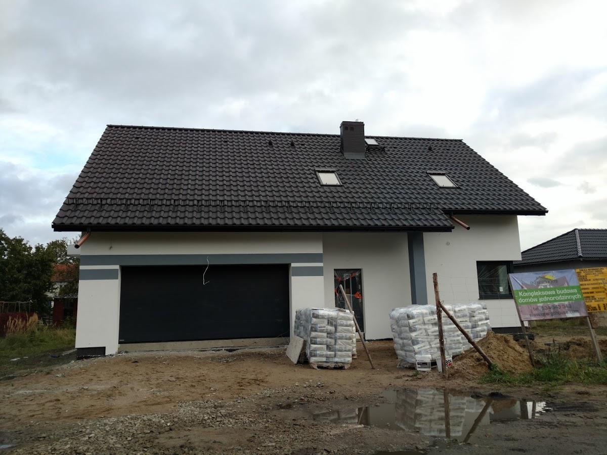 Nowo wybudowany dom jednorodzinny z ciemną dachówką i szarą stolarką, widoczne worki z materiałami budowlanymi na posesji, po deszczu.