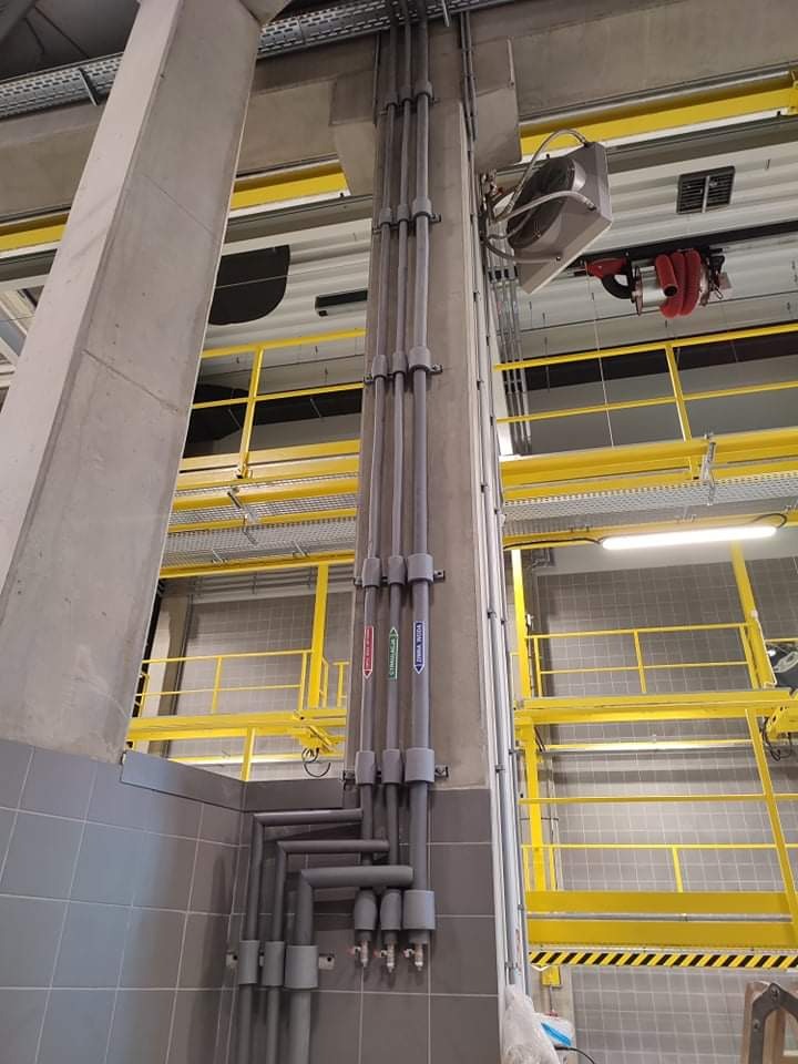 Szare rury instalacyjne prowadzone wzdłuż betonowego filaru wewnątrz budynku przemysłowego, z widocznymi etykietami 'zimna woda' i 'ciepła woda', żółte metalowe barierki w tle.