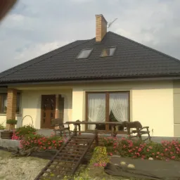 Jednorodzinny dom z ciemną dachówką, ceglanym kominem, ocieploną elewacją w odcieniach beżu i drewnianymi elementami dekoracyjnymi, otoczony rabatami z czerwonymi kwiatami.