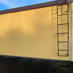Żółta elewacja budynku z przymocowaną metalową drabiną, widoczny fragment dachu pokrytego papą i fragment balustrady.