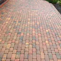 Ułożona kostka brukowa w odcieniach czerwieni, brązu i szarości, widok z perspektywy lekko z góry, z widocznym fragmentem budynku i zieleni w tle.