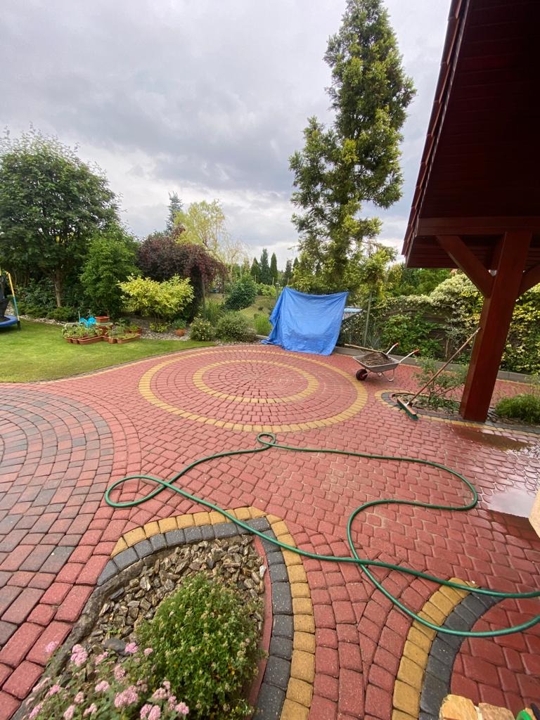 Czerwona kostka brukowa ułożona w dekoracyjne wzory na posesji w Pile, z zielonym wężem ogrodowym i widocznym fragmentem ogrodu z drzewami i krzewami w tle.