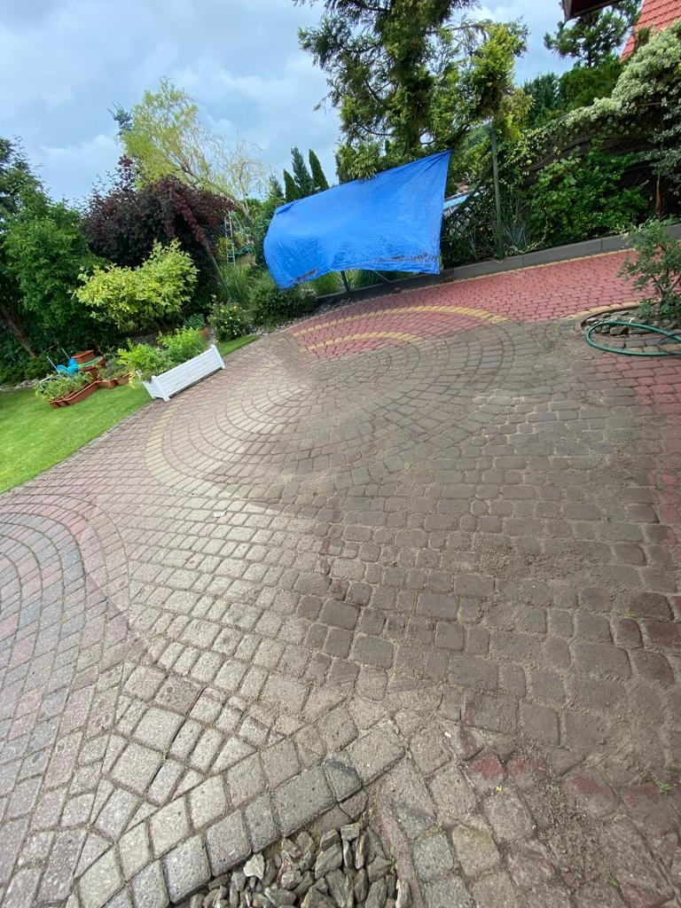 Umyta i brudna część kostki brukowej na podjeździe, z niebieską plandeką w tle i zielonym trawnikiem po lewej stronie.